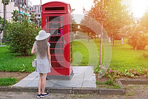 Tourist girl in a white hat near a British telephone booth. Red vintage british telephone box on street in park on a sunny day