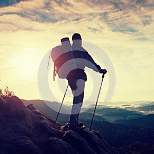 Tourist guide with pole in hand. Hiker with sporty backpack stand on rocky view point above misty valley. Sunny spring daybreak