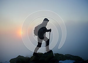 Tourist hiker man with backpack on rocky mountain on misty blue sky and raising sun background.