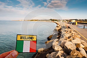 Tourist holding badge with sign Ireland and Irish flag in focus. Silverstrand beach at high tide out of focus. Blue cloudy sky.