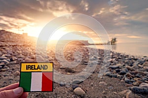 Tourist holding badge with sign Ireland and National flag . Salthill beach and Blackrock diving board out of focus. Sun rise time
