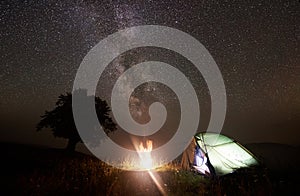 Tourist illuminated tent near big tree, camping in mountains at night under starry sky