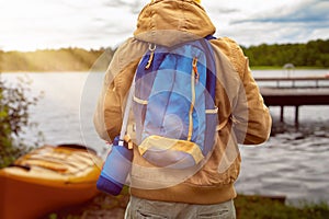 Tourist in a jacket and with a backpack on a journey against the backdrop of a kayak, forest and lake on a sunny day