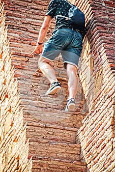 Tourist in jeans shorts and teeshirt with small backpack climbs steep narrow steps in ancient ruin - cropped photo