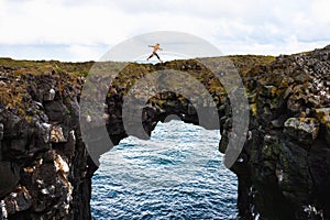 Tourist jumps over a natural rock bridge in Arnarstapi, Iceland photo