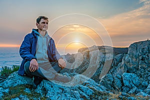 Tourist man in a blue jacket sits on a rock high in the mountains, above the clouds and enjoys the sunset Copy space. The concept
