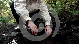 Tourist man squatting in front of a mountain river in dense forest in a sunny spring day. Footage. Man in white jacket