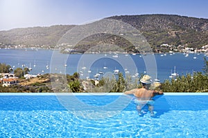 A tourist man in a swimming pool enjoys the view of the sailing boats in a bay at Poros island