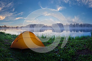 Tourist orange tent on green grass of foggy lake at sunrise