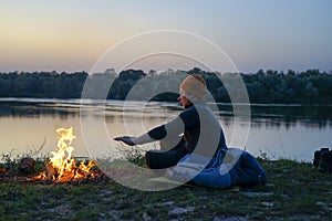 A tourist pours tea from a thermos on the river bank. The guy sits on a sleeping bag, and his things lie nearby. A tourist on a