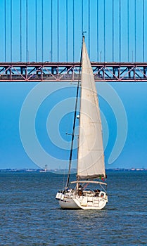 Tourist recreation sailboat sailing on the red steel 25 de Abril Suspension Bridge over the Tagus River in the city of Lisbon,