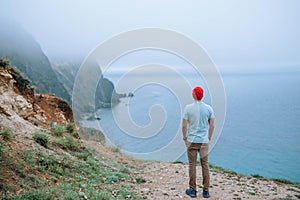 Tourist in a red cap stands on a cliff overlooking the sea.