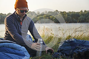 A tourist is resting on the bank of the river. He is in a sleeping bag.