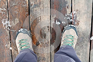 Tourist`s feet shod in pair of sport trekking boots on the old brown wooden boards surface outdoors in spring day top view