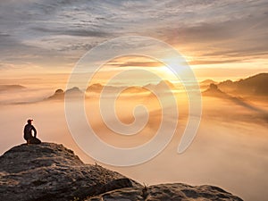 Tourist sit on peak of sandstone rock and watching into colorful mist and fog in morning valley. Sad man