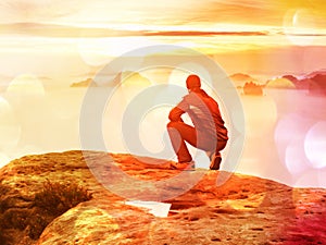 Tourist sit on peak of sandstone rock and watching into colorful mist and fog in morning valley. Sad man. Man sit. Man in jeans.