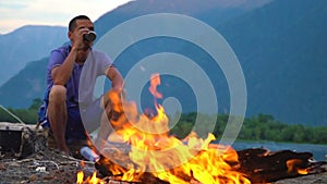 A tourist sits by the fire on the shore of a mountain lake.