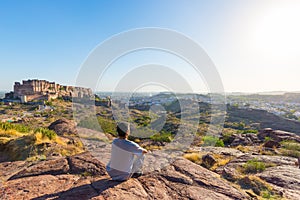 Tourist sitting on rock and looking at expansive view of Jodhpur fort from above, perched on top dominating the blue town. Travel