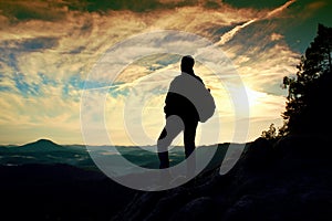 Tourist with sporty backpack stand on rocky view point and watching into deep misty valley bellow. Sunny spring daybreak in rocky