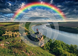 Tourist standing on the edge of the cliff and looking at the rainbow. Rainbow over the river