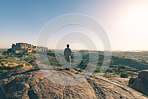 Tourist standing on rock and looking at expansive view of Jodhpur fort from above, perched on top dominating the blue town. Travel