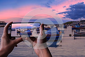 Tourist taking photo of traditional Maltese boats in Marsaxlokk harbor during sunset, Malta