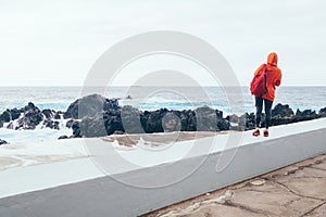 Tourist woman with backpack enjoying the power of ocean looking at waves surf at Porto Moniz natural pools, Madeira island,