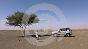 Tourist woman in dress walking away car 4x4 and beautiful tree alone in a desert landscape with sand dunes in background