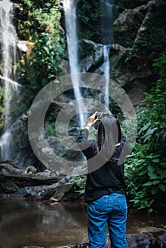 Tourist Woman is Enjoying While Photographing Waterfall on Travel Vacation, Rear View of Asian Woman Relaxing Outdoor Adventure