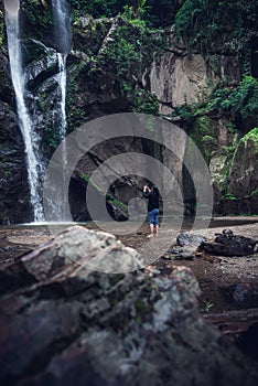 Tourist Woman is Enjoying While Photographing Waterfall on Travel Vacation, Rear View of Asian Woman Relaxing Outdoor Adventure