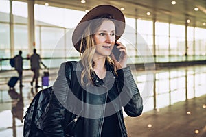 Tourist woman in hat and with backpack is standing at airport and talking on cell phone.Girl stands, uses digital gadget