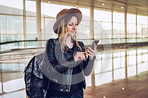 Tourist woman in hat, with backpack stands at airport, uses smartphone. Girl checks email, chatting, browsing internet.