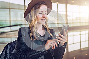 Tourist woman in hat, with backpack stands at airport, uses smartphone. Girl checks email, chatting, browsing internet.