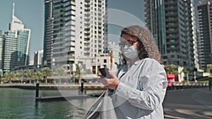 Tourist woman in medical face mask browsing phone walking on waterfront in city.