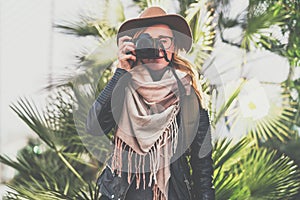 Tourist woman photographer in hat and with backpack stands outdoor on background of green plants and takes pictures.