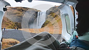 Tourist woman sit hold cup enjoy nature views with open door inside rented campervan. Skogafoss waterfall, Iceland. Travel solo