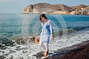 Tourist woman walking on coastline of Red beach Santorini island Greece barefooted. Traveling and summer vacation