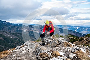 A tourist in a yellow cap and a red down jacket sits on a rock and puts crampons on his shoes.
