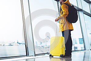 Tourist with yellow suitcase backpack is standing at airport on background large window, traveler man waiting in departure lounge