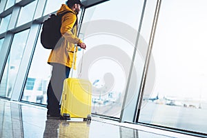 Tourist with yellow suitcase backpack is standing at airport on background large window, traveler person waiting in departure loun