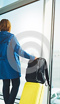 Tourist with yellow suitcase backpack is standing at airport on background large window, traveler woman waiting in departure loung