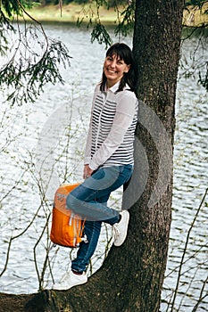 Tourist young smiling girl with orange backpack in hands standing near to big tree on bank of mountain lake surrounded by forest
