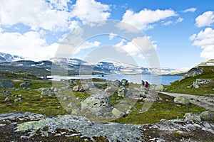 Tourists hiking towards dam of Lake Finsevatnet, snowy mountains and glacier Hardangerjokulen in Finse, Norway