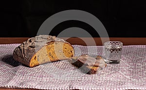 On a towel, bread, one pieces cut off and a short glass of vodka on a black background