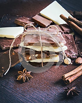 A tower of chocolate bars wrapped like a chocolate present . Various chocolate pieces, spices,cocoa powder and nuts over dark wood