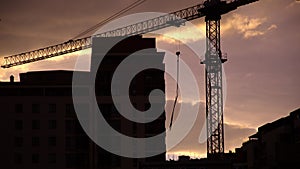 Tower crane stands still end of work day against clouded purple sky time lapse. Modern city buildings background under