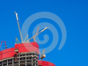 Tower cranes atop an unknown unfinished skyscraper against a clear blue sky