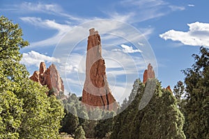 Towering red rock formations jut out of the ground to a height of 300 ft against a dramatic sky and surrounded by evergreen trees