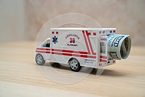 A toy ambulance on a wooden table with a rolled up one hundred American dollar bill in the back. High prices for ambulance