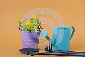Toy blue watering can, purple bucket with yellow-green sprigs, shovel and rake on an orange background